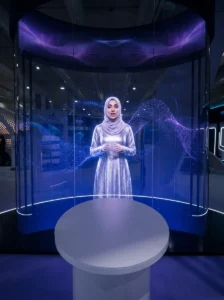 A life-size AI hologram host presenting inside a curved transparent LED booth at an exhibition